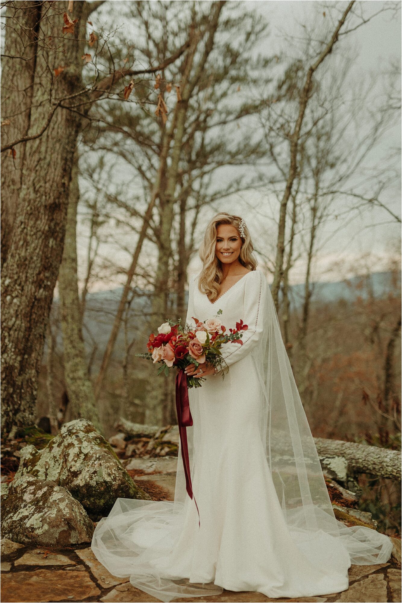 bride standing in woods wearing long white edgy gown with sleeves and sheer cape